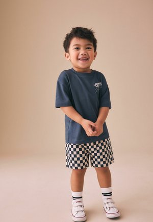 Lächelnder Kleinkindjunge steht mit verschränkten Händen da, trägt ein dunkelblaues T-Shirt, schwarz-weiß karierte Shorts, weiße Socken und Turnschuhe vor beigem Hintergrund.