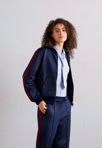 Donna con capelli ricci, vestita con una tuta navy con strisce rosse e una camicia azzurra, in piedi con una mano in tasca contro un muro semplice.