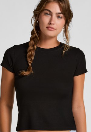 Jonge vrouw met lichtbruin gevlochten haar, die kleine ringoorbellen draagt en een zwart, aansluitend T-shirt met korte mouwen, op een neutrale achtergrond.