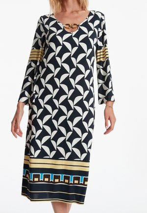 Kvinde iført knælængde sort kjole med hvidt bladlignende mønster, guldkædedetalje ved halsudskæringen, gyldne striber på ærmerne og geometrisk kant ved sømmen.