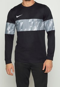 Muž v černém dlouhém rukávu Nike trička s reflexním šedým geometrickým vzorem přes hruď a horní část paží, doplněný černými kalhotami.