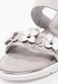 Sandale en daim gris avec des accents de fleurs blanches et des détails de perles argentées sur la sangle, dotée d'une semelle plate avec une bordure métallique.
