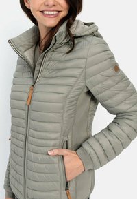 Veste matelassée en gris doux avec un col montant et une capuche. Dotée de poches zippées et de poignets côtelés, avec une étiquette en cuir.