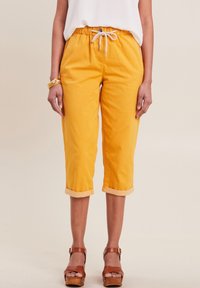 Gele cropped broek van katoen met een elastische tailleband en koord. Opgerolde zoom en zakken aanwezig. Bruine sandalen met houten zool zichtbaar.
