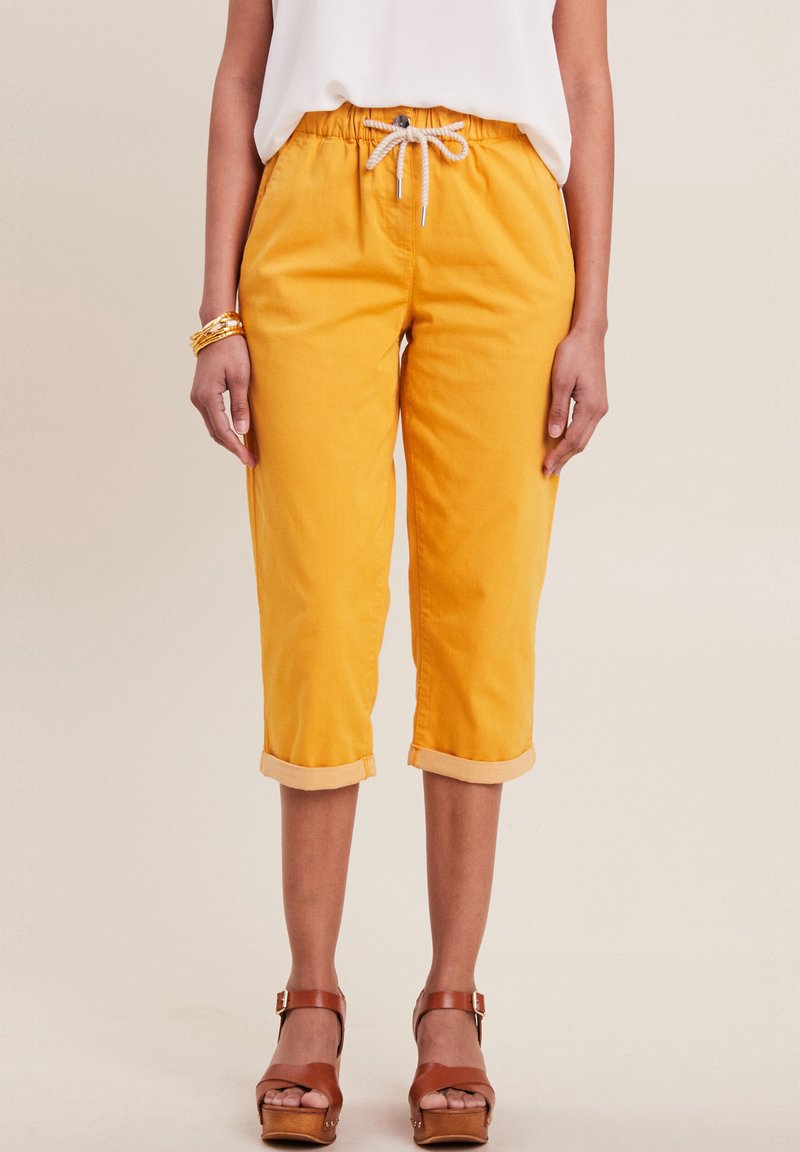 Gele cropped broek van katoen met een elastische tailleband en koord. Opgerolde zoom en zakken aanwezig. Bruine sandalen met houten zool zichtbaar.