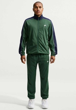 Mężczyzna stoi, ubrany w ciemnozielony dres Nike z granatowymi paskami i białe trampki, zwrócony przodem na jednolitym tle.