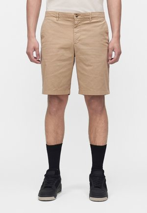 Homme portant un short beige arrivant au genou, des chaussettes noires à tige et des baskets noires, debout devant un fond blanc uni.