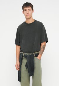 Tamno siva oversized majica s okruglim izrezom, uparena sa svijetlozelenim hlačama. Tamno pleteni materijal omotan oko struka za dodatni detalj.