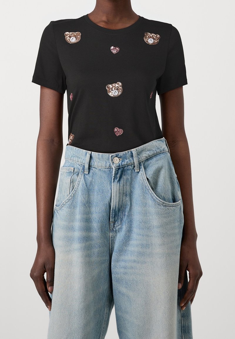 T-shirt noir à manches courtes avec des motifs métalliques d'ours et de cœur. Associé à un jean en denim bleu clair, taille haute, coupe décontractée.