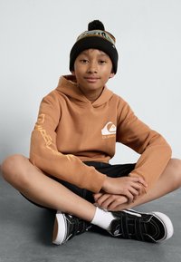 Bruine hoodie met logo, zwarte gebreide muts met een patroonband, zwarte shorts en zwarte schoenen met klittenbandsluitingen; zittend op een grijze ondergrond.
