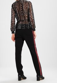 Persona che indossa una blusa nera trasparente a pois e pantaloni neri con strisce laterali rosse, in piedi su uno sfondo chiaro e semplice.