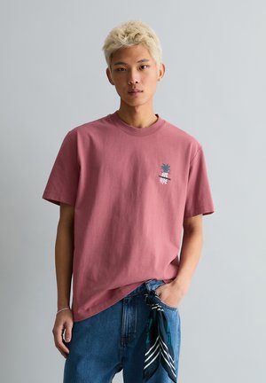 Young person with short bleached hair wearing a dusty rose T-shirt with small pineapple graphic and blue jeans with a dark scarf tied to the pocket.