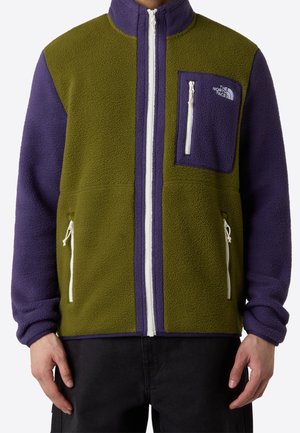 Persoon met een fleecejack met een olijfgroen lijf, paarse mouwen, borstzak en witte ritsdetails.