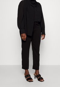 Svart långärmad skjorta med knappar kombinerad med skräddarsydda svarta byxor. Skorna är svarta, plisserade mule-sandaler med stilettklack.