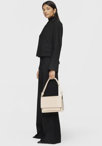 Borsa a mano in pelle beige con logo "TOUS" in rilievo, forma strutturata, manico a nodo corto, abbinata a un blazer nero e pantaloni a gamba larga.