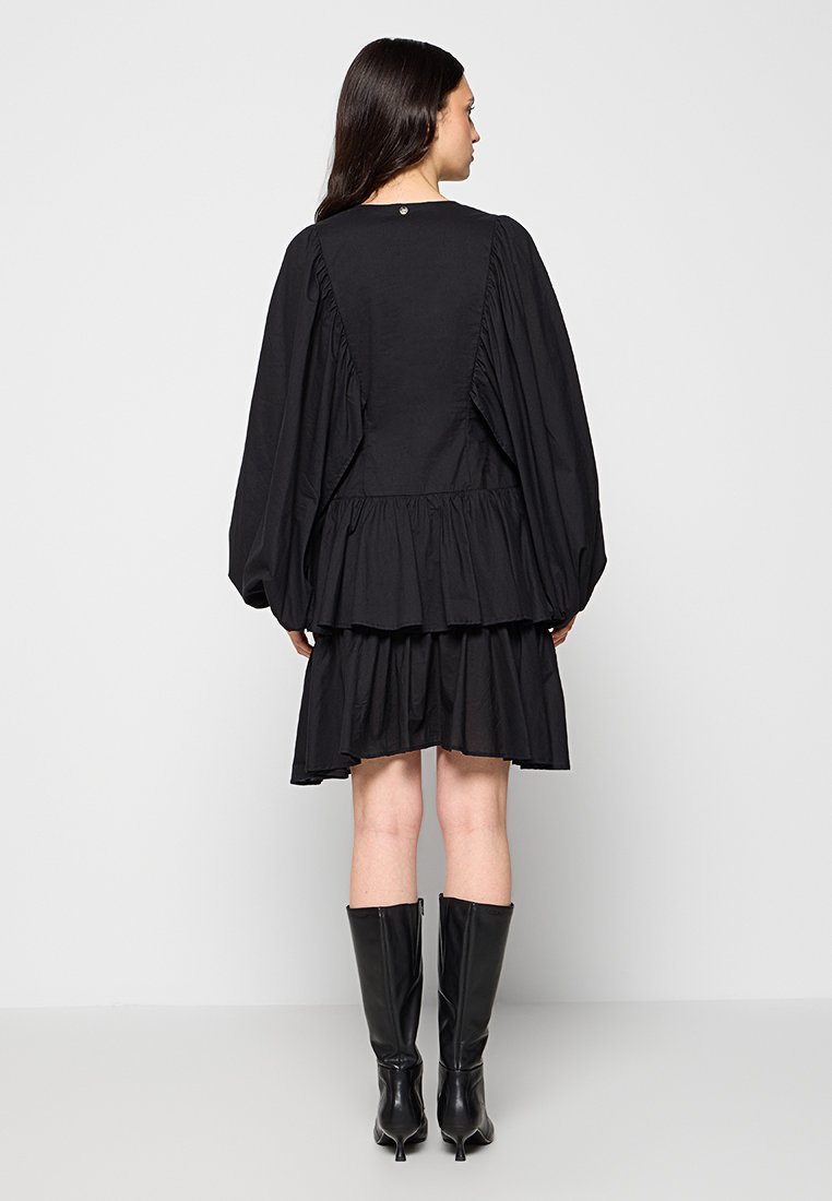 Schwarzes Kleid mit einem geschichteten Rüschensaum, voluminösen Ballonärmeln und einem Knopfdetail hinten. Glattes Material mit matter Oberfläche.
