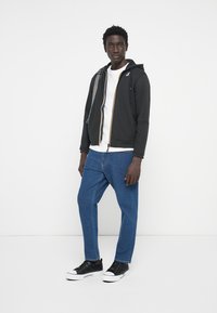 Svart zip-up hettegenser med fargerik glidelås, hvit t-skjorte, blå denimbukser og svarte sneakers, med kontrasterende teksturer og uformell design.