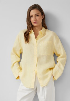 Frau mit langen braunen Haaren, die ein locker sitzendes, hellgelbes Hemd mit Knöpfen und weiße Hosen trägt, steht vor einem einfarbigen hellgrauen Hintergrund.