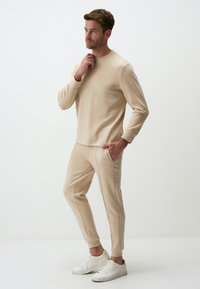 Beige cotton sweatshirt with long sleeves and round neck, paired with matching tapered pants featuring side pockets and trim accents. White sneakers.