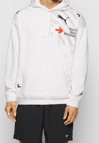 Bílá kapuce s tečkami a přední kapsou, kapuce se šňůrkami, šedé grafiky a červený text "REDUCE REUSE REGENERATE." V kombinaci s černými šortkami.