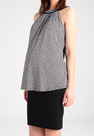 Kvinde iført ærmeløs halterneck-top i sort og hvid ternet mønster samt sort knælang pencil skirt, stående mod en ensfarvet baggrund.