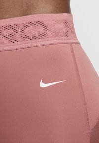 Nike tajice od mekanog ružičastog materijala s teksturiranim pojasom. Imaju bijeli Nike logo na bedru. Glatka površina, prilagođeni dizajn.
