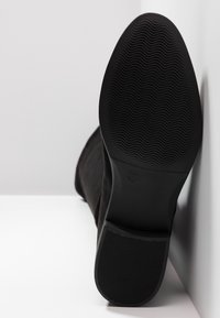 Black suede boot showing textured rubber sole with wavy patterns, size marked as 37, angled against a light background.
