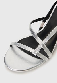 Sandalo piatto metallico argento con cinturini incrociati, texture liscia e design a punta arrotondata. La soletta nera contrasta con la parte superiore.