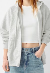 Felpa leggera grigia con zip, dalla finitura morbida e testurizzata, dotata di tasca a marsupio e polsini a costine, indossata sopra una maglietta bianca aderente.