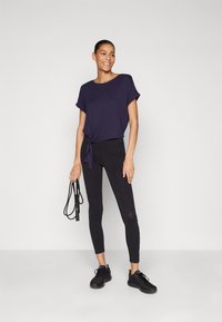Marineblå t-skjorte med knyttet frontdetalj, sammen med tettsittende svarte leggings. Modellen holder et svart hoppetau og har på seg svarte treningssko.