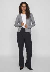Grijze gebreide cardigan met open voorkant, ribbelmanchetten, gelaagd over een witte top; gecombineerd met zwarte wijde broek en zwarte hakken.