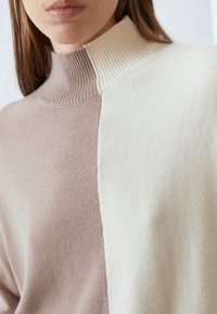 Femme portant un pull en maille douce à col roulé avec un design bicolore beige et crème, présenté des épaules aux lèvres.