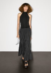 Top nero con scollatura a halter abbinato a una gonna maxi nera fluente e a strati. La modella tiene una pochette bianca. Tessuto morbido, design minimalista.