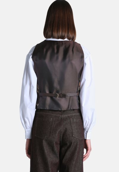Femme aux cheveux bruns et raides portant un gilet formel noir sur une chemise blanche à manches longues et un pantalon foncé taille haute, vue de dos.