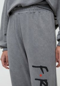 Person iført grå joggebukser og matchende sweatshirts med sort og rød abstrakt tekstdesign på buksebenet.
