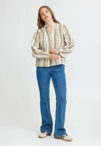 Kvinde står iført en beige stribet bluse, blå jeans og beige sneakers med sorte striber mod en ensfarvet hvid baggrund.