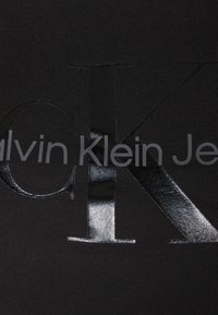 Melnas drēbes ar ribotu tekstūru, centrā izceļas izteiksmīgs, spīdīgs melns "CK" logo, kas norāda "Calvin Klein Jeans" pelēkā krāsā.