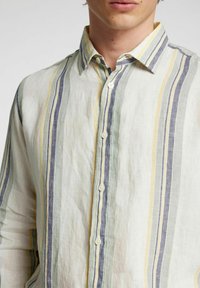 Camicia di lino con righe verticali in giallo pallido, verde e blu. Colletto classico, chiusura con bottoni e texture morbida.