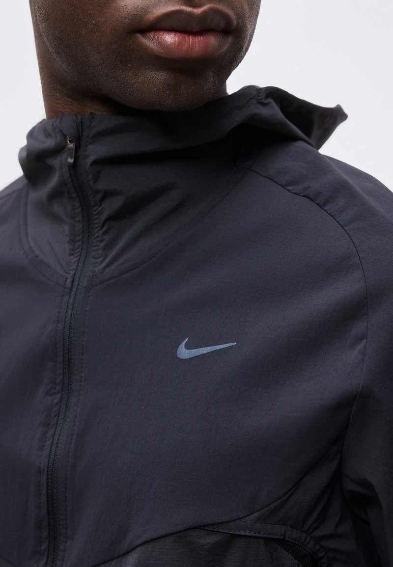 Primer plano de una persona que lleva una chaqueta negra con cremallera de Nike con el logo en el pecho izquierdo, mostrando la parte inferior del rostro y el cuello.
