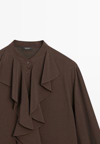 Blouse marron à boutons avec un détail à volants sur le devant et un col rond, présentée sur un fond blanc.