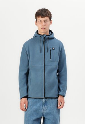Gråblå hettejakke med glidelås på brystlommen, teksturert stoff og svarte glidelåsdetaljer. Brukes sammen med lyseblå denimjeans.