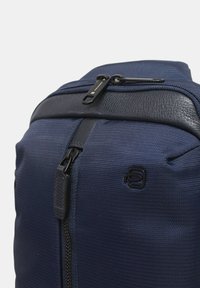 Mochila texturizada azul marino con cremalleras negras y detalles de cuero, que cuenta con dos tiradores de cremallera y un pequeño logo negro en el bolsillo delantero.