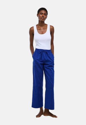 Femme debout pieds nus portant un débardeur blanc et un pantalon bleu ample, les mains dans les poches, devant un fond uni.