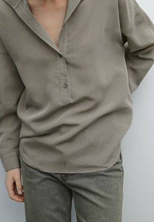Person iført en løs, taupefarvet, tyndstribet bluse delvist knappet, kombineret med grå denimjeans, stående mod en ensfarvet baggrund.