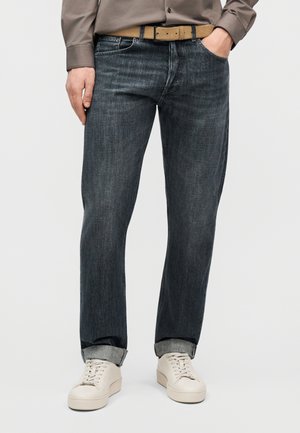 Άνδρας που φοράει σκούρα γκρι τζιν με γυρισμένα μπατζάκια, μπεζ ζώνη, πουκάμισο τωπ και λευκά αθλητικά παπούτσια, στέκεται με το ένα χέρι στην τσέπη.