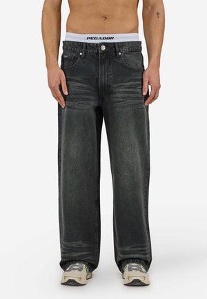Mann trägt locker sitzende, ausgewaschene schwarze Jeans, weiße Unterwäsche mit Bund und beige Sneaker, steht mit entspannten Armen neben dem Körper.