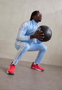 Survêtement de sport bleu clair avec des rayures blanches, présentant une conception ajustée. Un ballon de médecine noir tenu dans les mains, des baskets roses avec des accents.