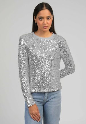 Femme portant un top argenté à sequins à manches longues et un jean bleu clair, debout devant un fond gris uni, une main derrière le dos.