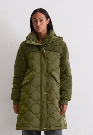 Manteau long matelassé vert olive avec une partie supérieure texturée et duveteuse, deux poches avant et une capuche amovible avec des fermetures éclair.