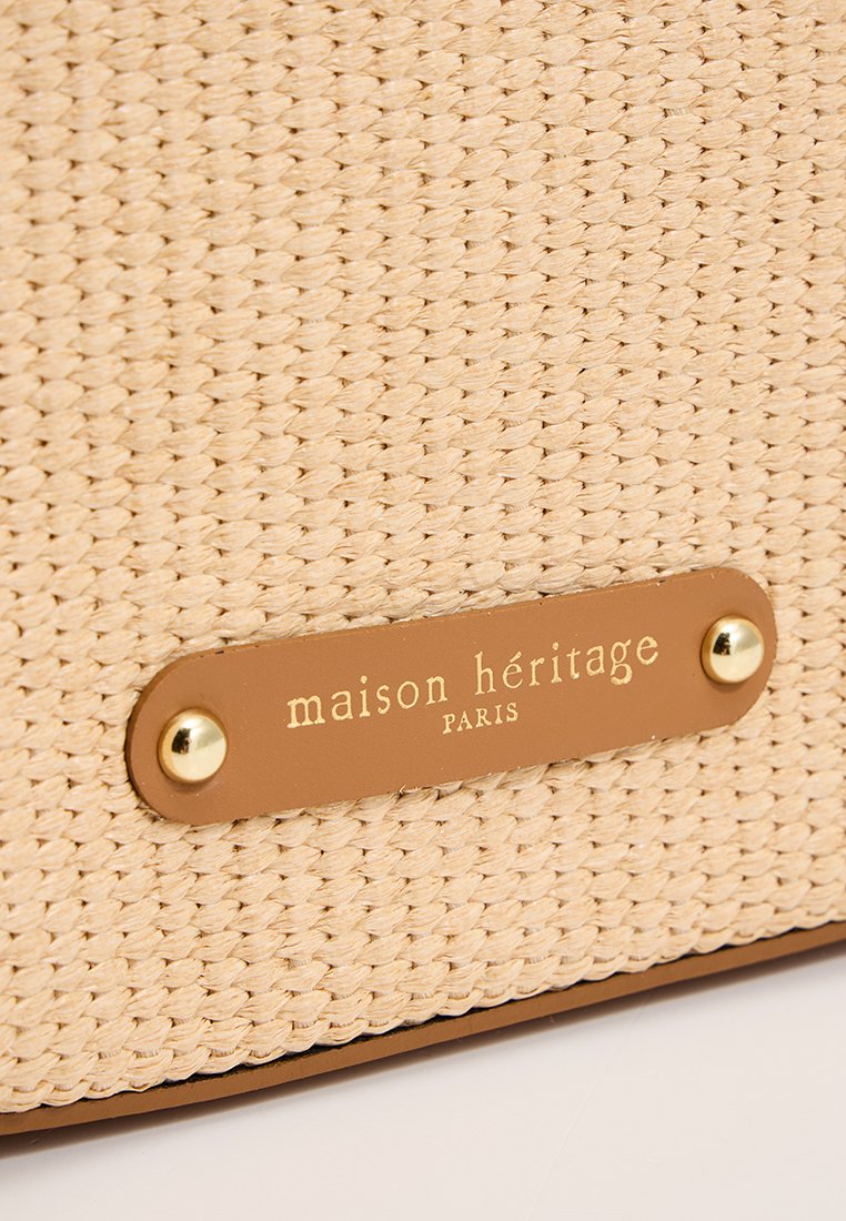 Beige geweven strooien tas met een gestructureerd oppervlak, voorzien van een bruine leren label met gouden letters en hardware accenten.